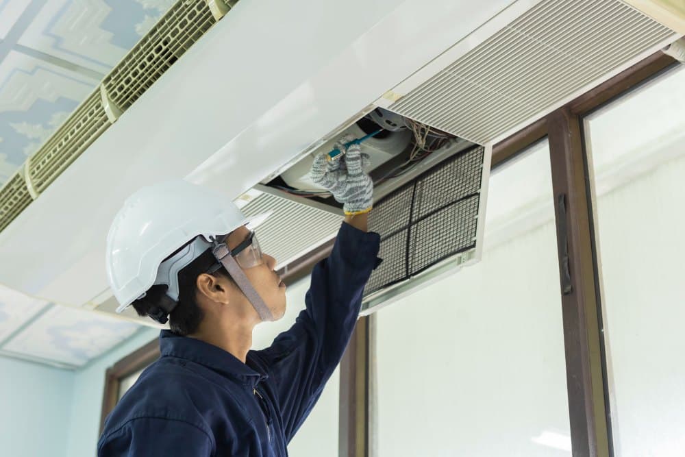 A technician in a hard hat and safety glasses opening the panel of a ceiling cassette to reach its filter.