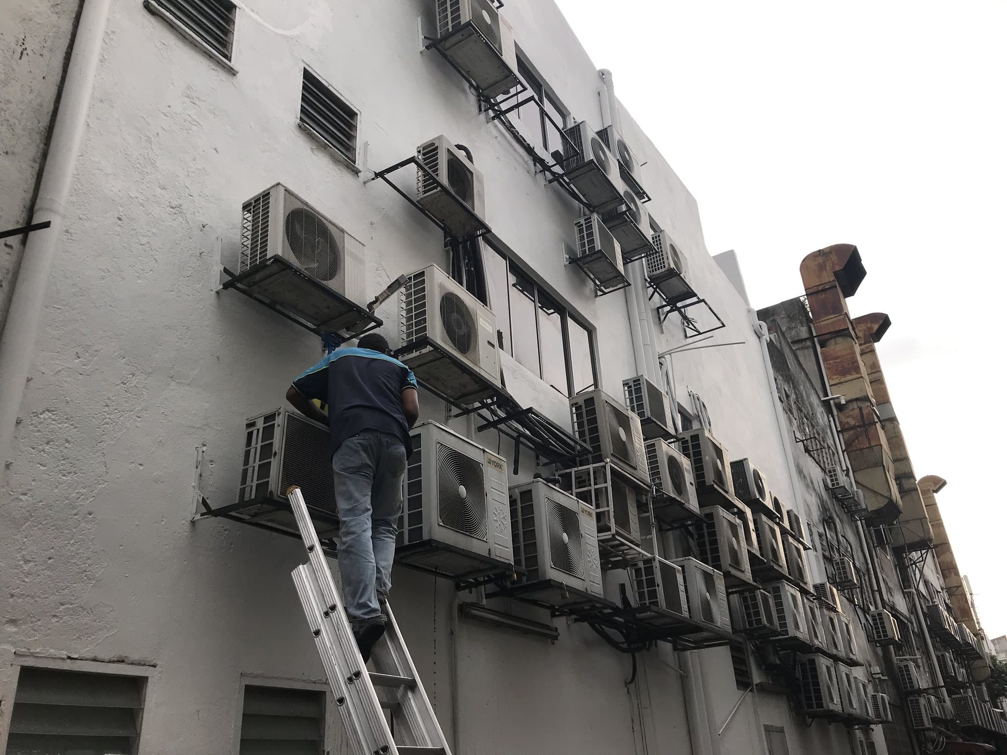 A technician on a ladder reaching a bank of wall-mounted condensing units stacked several storeys up a building facade.
