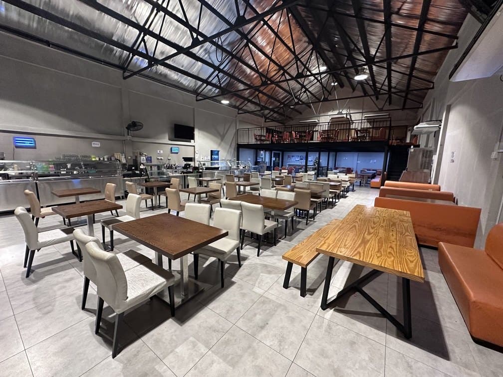 The hospital cafeteria seen from the servery end, tables and banquettes under an exposed steel roof.