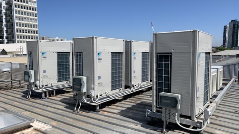 Four Daikin VRV condensing units standing in a row on a metal roof deck, refrigerant pipework and isolators running between them.