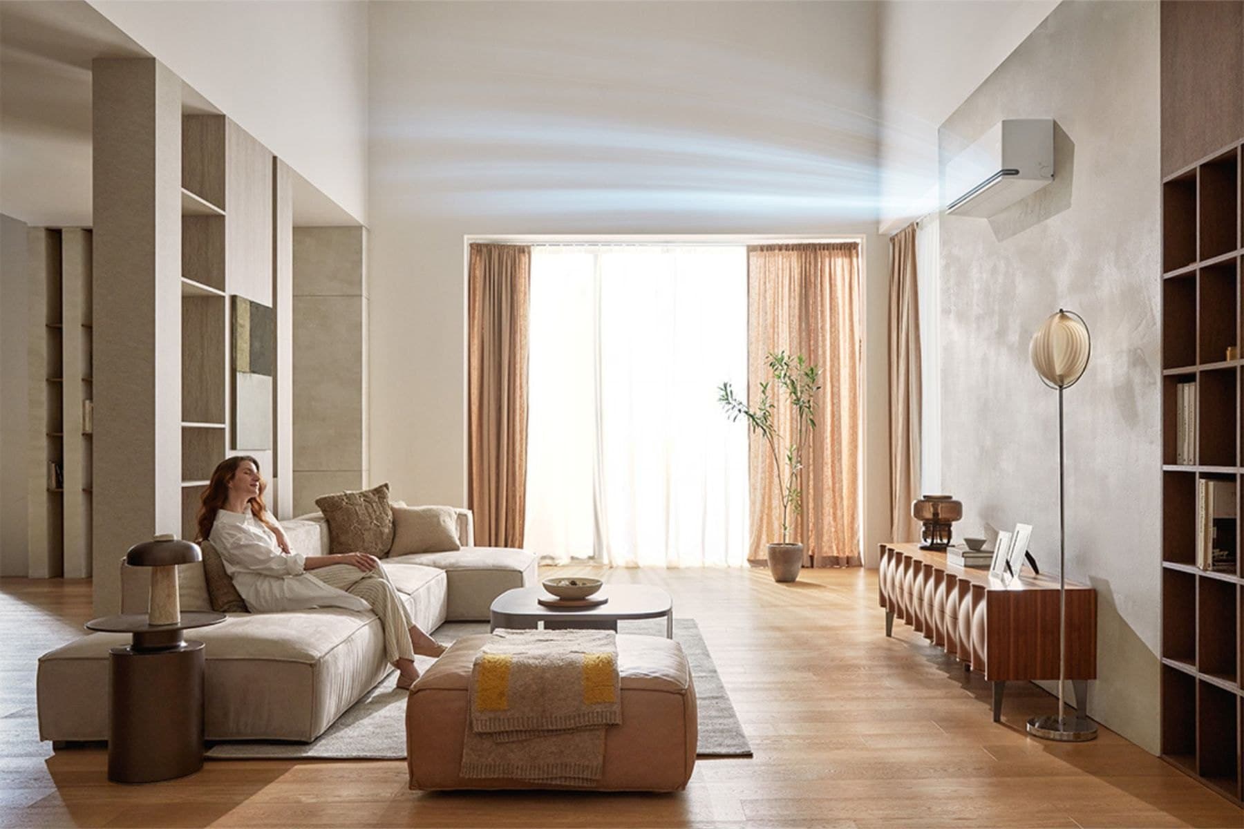A double-height living room with a wall-mounted indoor unit set high on the far wall, an illustrated sweep showing conditioned air carried across the ceiling toward the seating.