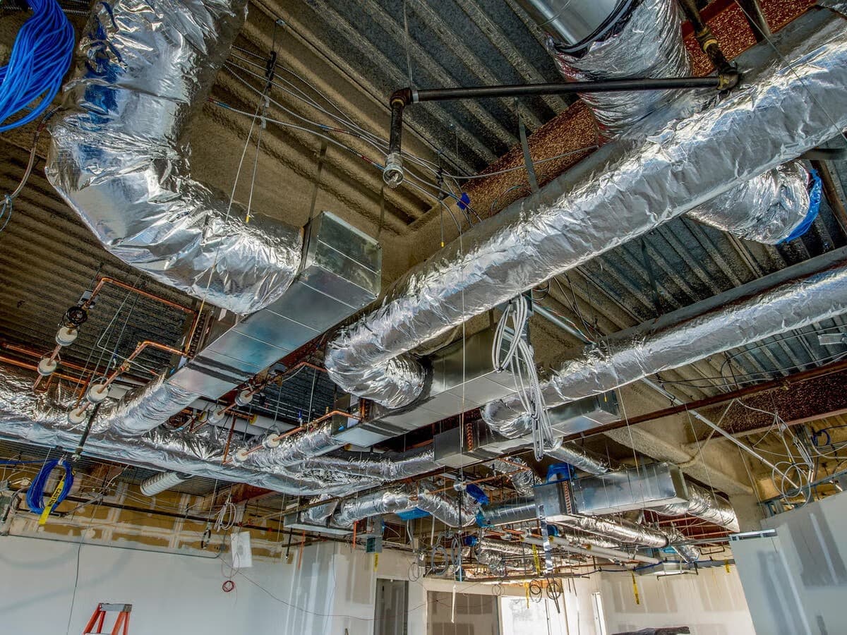 A ceiling void under construction with insulated flexible ducts, rectangular sheet-metal trunking, copper pipework and cabling running between the structural beams.