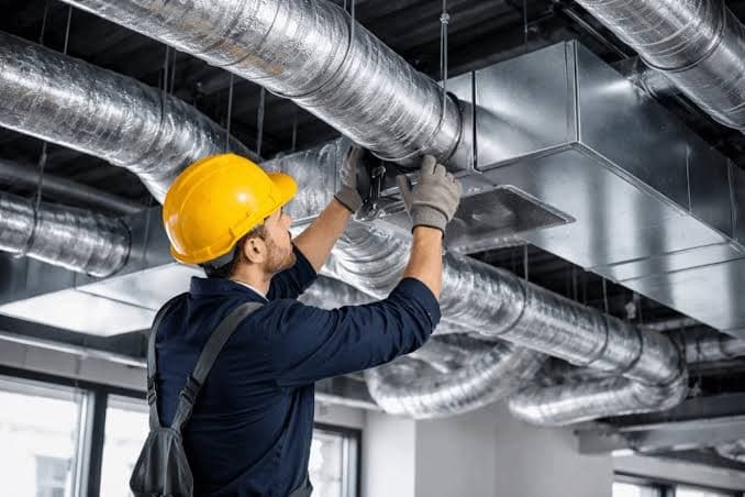 A technician in a hard hat working at the connection between a spiral duct and a ceiling diffuser box.
